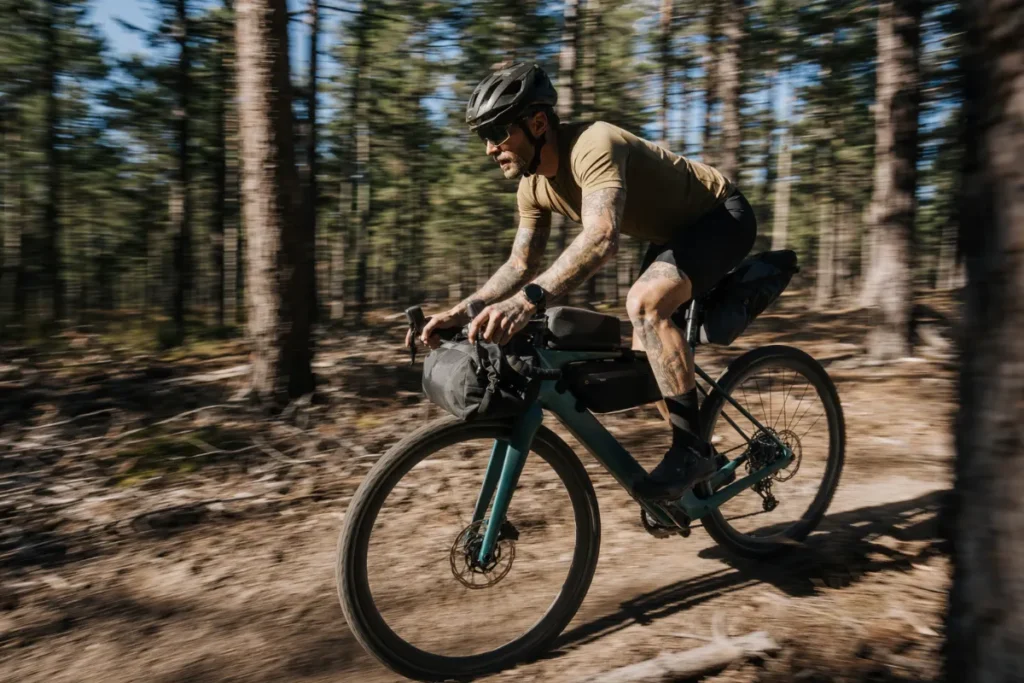 Kolesar na makadamskem kolesu z nepremočljivimi bikepacking torbami vozi po gozdni enoslednici.